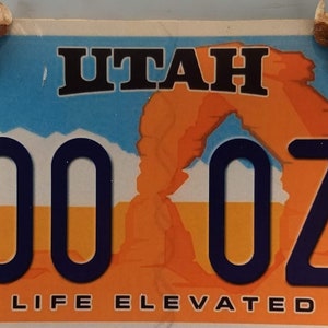 Op de afbeelding: Een blauw en oranje Utah kenteken met de tekst "ZOO 0ZZ" en de slogan "LIFE ELEVATED". De plaat heeft een rode rotsboog en twee hertenkop ornamenten.