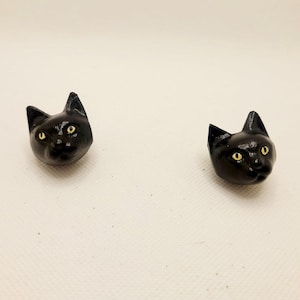 Puede incluir: Dos figuritas de cabeza de gato negras con ojos amarillos. Las cabezas de gato están mirando hacia adelante y están hechas de un material negro liso.