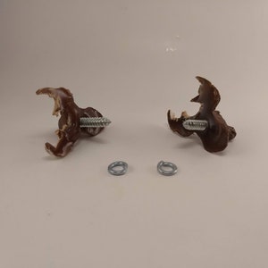 Op de afbeelding: Twee bruine plastic wandhaken met een schroef aan elk. Twee zilveren metalen ringen zijn ook inbegrepen.