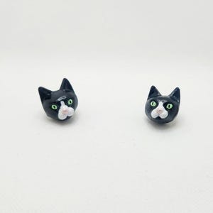 Puede incluir: Un par de pendientes de cara de gato negro y blanco con ojos verdes.