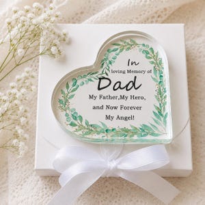 May include: A clear heart-shaped memorial plaque with the words "In loving Memory of Dad" and "My Father, My Hero, and Now Forever My Angel!" in black script. The plaque is decorated with green leaves and rests on a white box with a white ribbon.