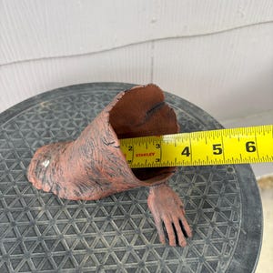 May include: A brown, textured, hollow, arm-shaped object with a hand extending from the base. A yellow Stanley measuring tape is positioned inside the opening, showing a diameter of approximately 3.5 inches across the interior of the opening.