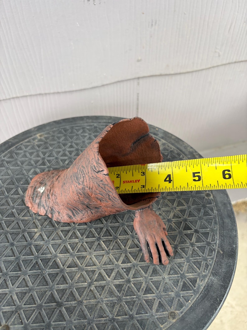 May include: A brown, textured, hollow, arm-shaped object with a hand extending from the base. A yellow Stanley measuring tape is positioned inside the opening, showing a diameter of approximately 3.5 inches across the interior of the opening.