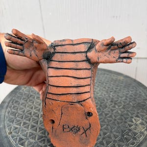 May include: A terracotta-colored ceramic sculpture featuring a textured, segmented body with two outstretched hand-like appendages. The base displays the hand-etched text "BOX" with a small circular symbol between the letters O and X.