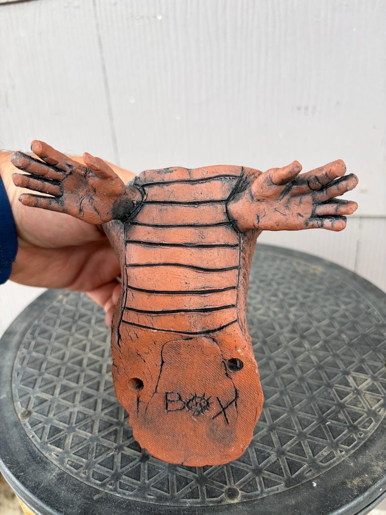 May include: A terracotta-colored ceramic sculpture featuring a textured, segmented body with two outstretched hand-like appendages. The base displays the hand-etched text "BOX" with a small circular symbol between the letters O and X.