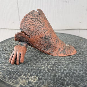 May include: A terracotta-colored ceramic sculpture depicting a hand emerging from a textured, sleeve-like form. The surface features a dark, weathered patina with intricate, vein-like detailing across the entire exterior of the object.