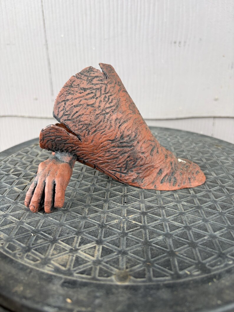 May include: A terracotta-colored ceramic sculpture depicting a hand emerging from a textured, sleeve-like form. The surface features a dark, weathered patina with intricate, vein-like detailing across the entire exterior of the object.