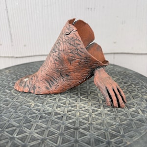 May include: A terracotta-colored ceramic sculpture featuring a textured, cone-shaped body with dark, etched markings and a single, realistic hand extending from the side. The object has an open top and a wide, flat base, showcasing a raw, earthy finish.