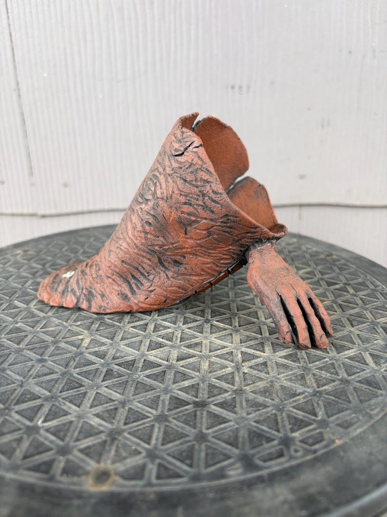 May include: A terracotta-colored ceramic sculpture featuring a textured, cone-shaped body with dark, etched markings and a single, realistic hand extending from the side. The object has an open top and a wide, flat base, showcasing a raw, earthy finish.