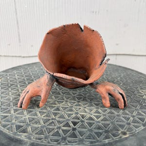 May include: A terracotta-colored ceramic vessel with a wide, flared, organic-shaped opening and two sculpted hand-like appendages extending from the sides. The surface has a textured, matte finish with dark, charred-looking accents along the edges.