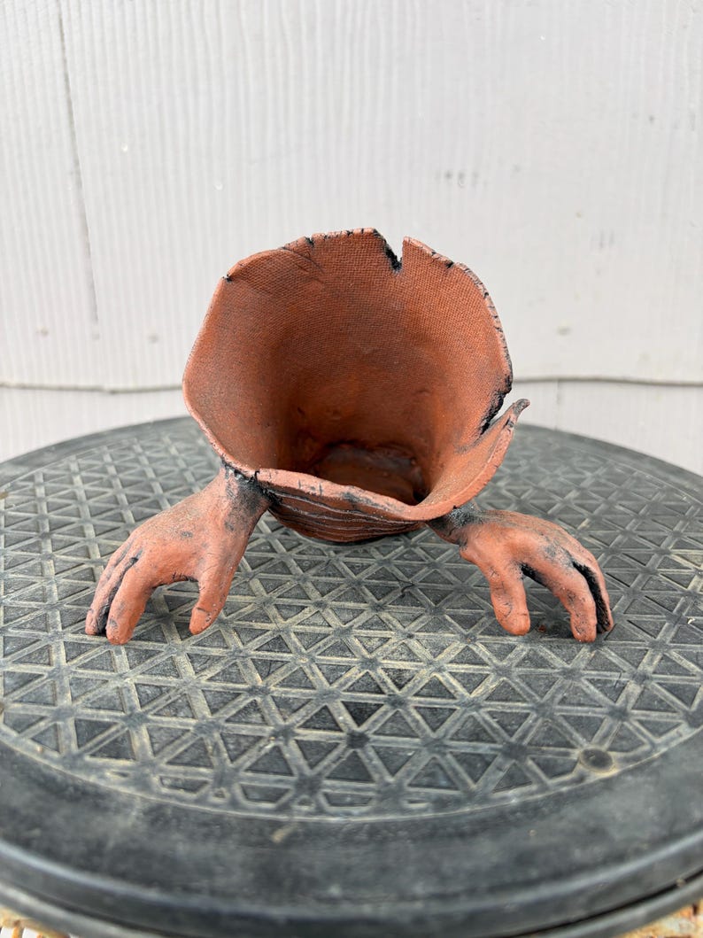 May include: A terracotta-colored ceramic vessel with a wide, flared, organic-shaped opening and two sculpted hand-like appendages extending from the sides. The surface has a textured, matte finish with dark, charred-looking accents along the edges.