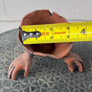 May include: A terracotta-colored vessel shaped like a hollow stump with two hand-like appendages extending from the base. A yellow Stanley measuring tape is positioned across the opening, showing a diameter of approximately 3.5 inches.