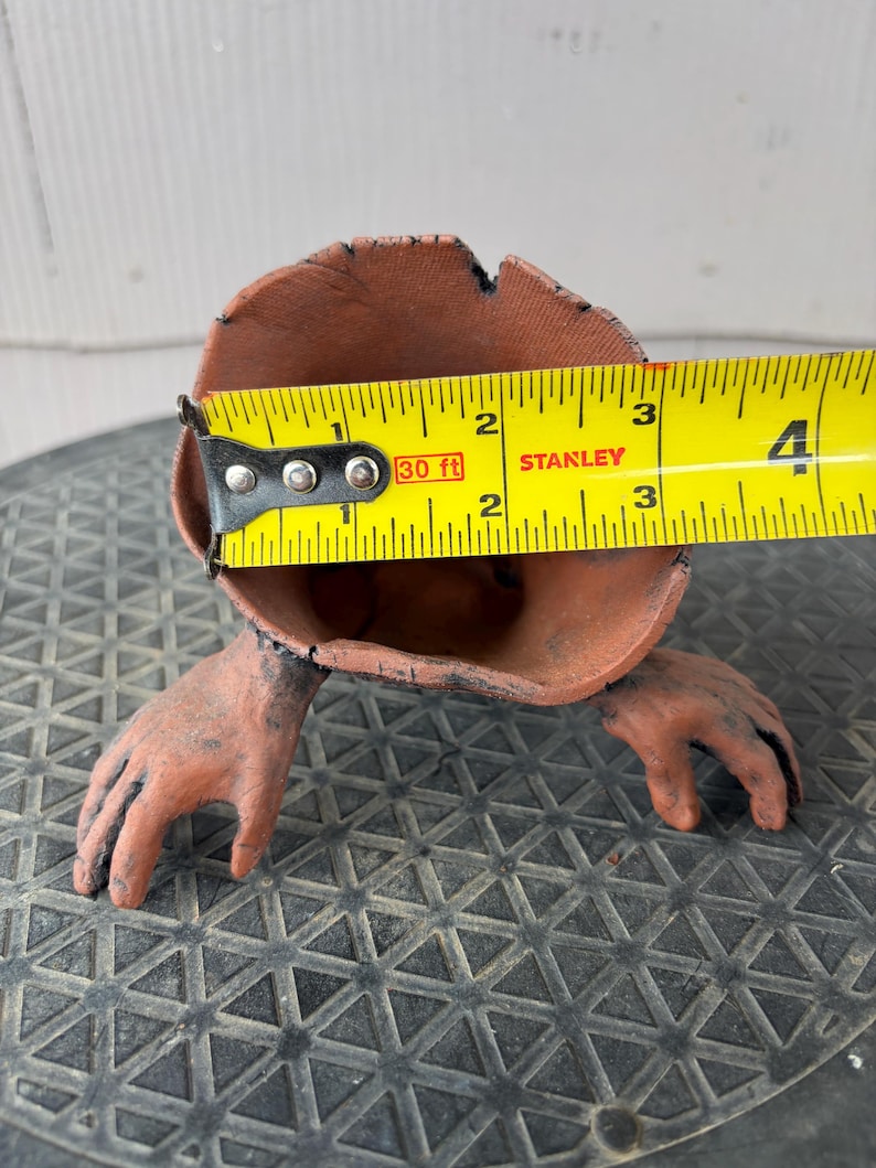 May include: A terracotta-colored vessel shaped like a hollow stump with two hand-like appendages extending from the base. A yellow Stanley measuring tape is positioned across the opening, showing a diameter of approximately 3.5 inches.