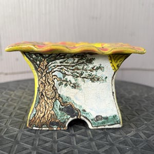 May include: A square ceramic planter with a flared, wavy rim featuring a yellow and pink pattern. The exterior sides are decorated with a hand-painted scene of a gnarled brown tree, green grass, and dark rocks against a white background.