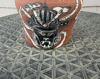 May include: A terracotta-colored clay pot featuring a hand-painted black and white graphic of a samurai helmet and mask. The design includes intricate details of the helmet's crest and the fierce expression of the mask, with white rope-like accents.