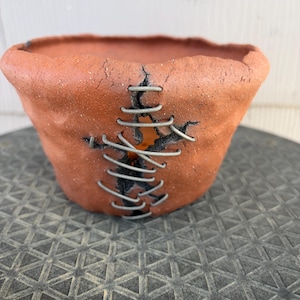 May include: A terracotta-colored ceramic bowl with a rough, uneven texture. The bowl features a vertical crack down the center that is held together by a series of grey metal staples, creating a stitched appearance across the exterior surface.