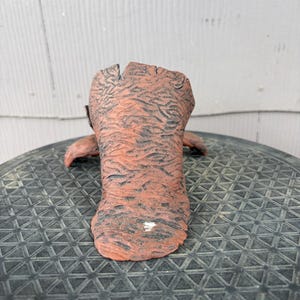 May include: A terracotta-colored ceramic vessel with a rough, textured surface resembling tree bark. The object features a wide, open top and a flared base, with dark, recessed grooves throughout the exterior and a small white mark near the bottom.
