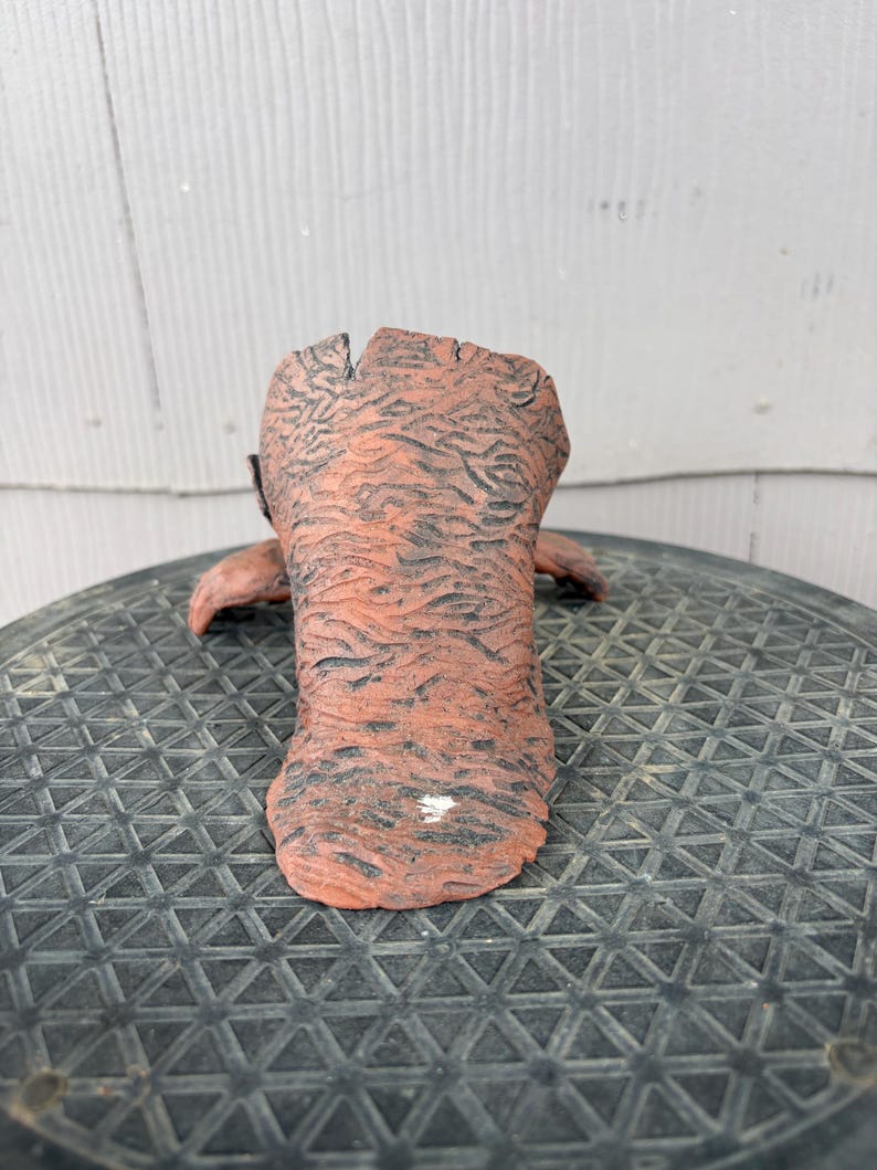 May include: A terracotta-colored ceramic vessel with a rough, textured surface resembling tree bark. The object features a wide, open top and a flared base, with dark, recessed grooves throughout the exterior and a small white mark near the bottom.