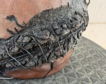 May include: A brown ceramic bowl featuring a textured, dark, charred-looking bark-like section attached to the side. The dark section is secured to the smooth brown clay surface with thin, dark metal wire wrapped in a crisscross pattern.