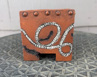 May include: A square terracotta-colored planter featuring a hand-painted white rope design that loops across the front surface. The top edge is decorated with five raised circular studs, and the base includes a cutout notch with dark distressed detailing.