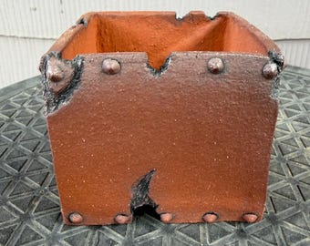 May include: A square terracotta planter with a rough, textured surface and dark, weathered edges. The exterior features small, raised, rivet-like details along the top and bottom rims, with a deep, dark, irregular notch cut into the center front.