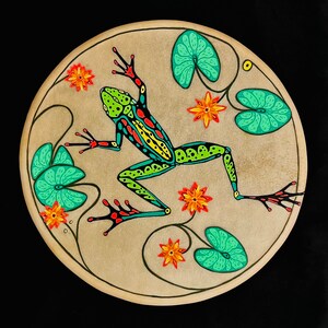Op de afbeelding: Een rond, beige canvas met een groene, gele en rode kikker erop geschilderd. De kikker wordt omringd door groene bladeren en oranje bloemen.