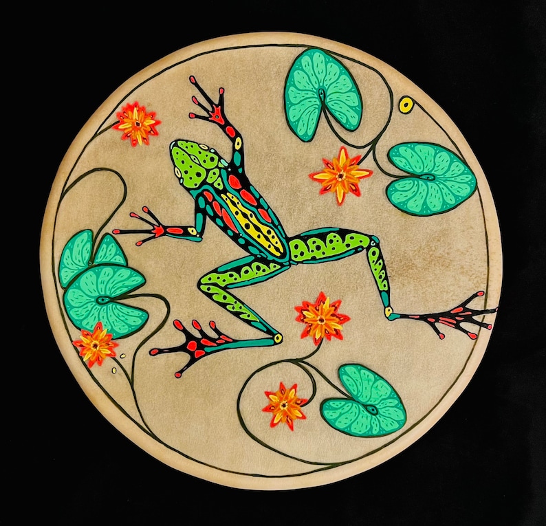 Op de afbeelding: Een rond, beige canvas met een groene, gele en rode kikker erop geschilderd. De kikker wordt omringd door groene bladeren en oranje bloemen.