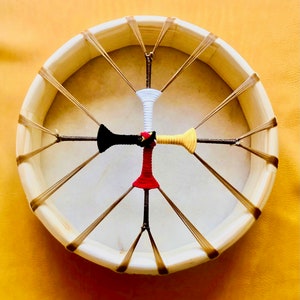 Op de afbeelding: Een houten raamtrommel met een wit leren vel. De trommel heeft bruin leren veters en een kruisvormig ontwerp in het midden. Het kruis is gemaakt van vier stokken die zijn omwikkeld met rood, wit, zwart en geel stof.