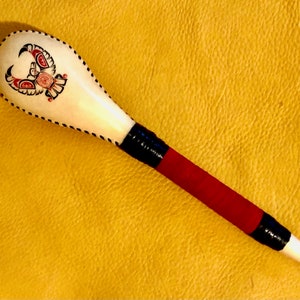May include: A white wooden spoon with a red, black and white design of a bird on the handle. The spoon is wrapped in red and black fabric.