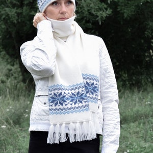 Puede incluir: Conjunto de gorro y bufanda de punto blanco con un patrón de copo de nieve azul. El gorro tiene un borde abatible y la bufanda tiene flecos.