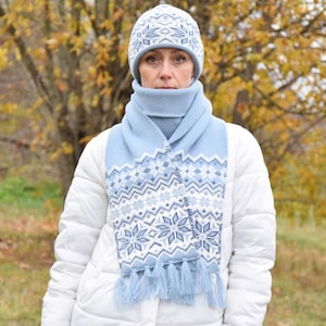 May include: A blue knitted hat and scarf set with a white snowflake pattern. The hat has a pompom on top and the scarf has tassels.