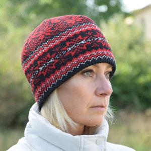 Può includere: Una donna indossa un berretto di lana rosso, nero e bianco con un motivo geometrico. Sta guardando da un lato e ha i capelli biondi.