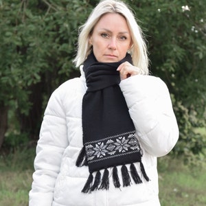 May include: A black knitted scarf with a white snowflake pattern and black tassels. The scarf is worn by a person with blonde hair.