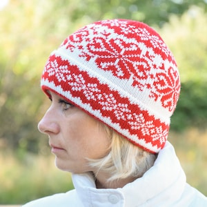 May include: A red and white knitted beanie with a snowflake pattern. The beanie is worn by a person with blonde hair.