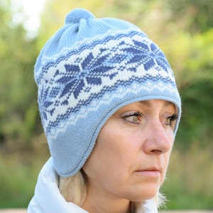 Può includere: Un cappello blu lavorato a maglia con un motivo a fiocco di neve bianco e blu. Il cappello ha le alette per le orecchie e un pon pon in cima.