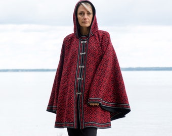 May include: A hooded cape in red and black with a geometric pattern. The cape features silver clasps down the front and black trim. The person is wearing black pants. The background is a body of water and a cloudy sky.