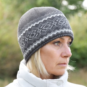 Può includere: Una donna indossa un berretto di lana grigio e bianco con un motivo geometrico. Il berretto è tirato giù sulla fronte e le copre le orecchie. Indossa una giacca bianca e ha i capelli biondi.