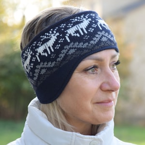 May include: A blue and white knitted ear warmer headband with a reindeer pattern. The headband is being worn by a person with blonde hair.