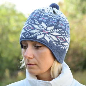 May include: A blue and white knitted hat with a pom pom on top. The hat has a snowflake pattern and ear flaps.