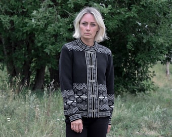 May include: A black knit cardigan featuring a white and grey geometric pattern along the collar, cuffs, and front placket. The garment includes a high neckline and is secured down the center with three silver-toned metal clasps.
