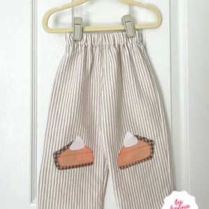 Puede incluir: Pantalones de rayas verticales en beige y blanco con cintura elástica. Cada pernera presenta un aplique de una porción de tarta de calabaza con una corteza a cuadros marrones, un relleno naranja y un copete de nata montada blanca.