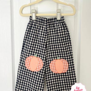 May include: Black and white gingham trousers featuring two orange and white chequered pumpkin appliqués on the knees. The trousers have an elasticated waistband and are displayed on a beige plastic hanger. A small white label reads by hanna june.