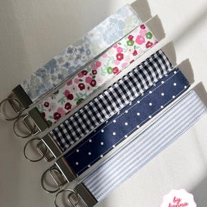 May include: Five fabric wristlet keychains with silver-tone metal hardware and split rings. The patterns include a light blue floral, a pink and green floral, a black and white gingham check, a navy blue with white polka dots, and a light blue pinstripe.