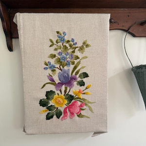 May include: A rectangular piece of light beige linen fabric featuring a central floral embroidery design. The embroidery displays a vertical arrangement of blue, purple, yellow, and pink flowers with green leaves and stems stitched onto the fabric.