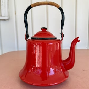 May include: A vibrant red enamel kettle with a black handle and spout. The handle is arched with a woven rattan grip. The kettle has a matching red lid with a black knob. The kettle is sitting on a pink surface.
