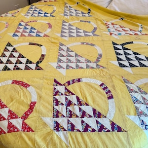 May include: A yellow quilt featuring a repeating basket pattern. Each basket is constructed from various colorful patterned fabric triangles and a solid white handle. The quilt is composed of multiple square blocks stitched together in a grid.