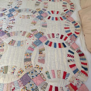 Op de afbeelding: Een quilt met een dubbel trouwringpatroon op een gebroken witte achtergrond, bestaande uit in elkaar grijpende ringen van diverse kleine lapjes stof met bloemen-, ruit- en stippenpatronen in rood, blauw, roze, geel en groen met zichtbaar stikwerk.