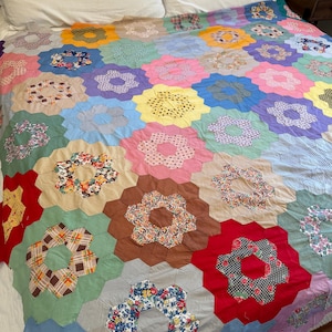 Op de afbeelding: Een handgemaakte patchwork quilt met een herhalend zeshoekig bloemenpatroon. De quilt is samengesteld uit talloze stoffen stukjes in diverse kleuren, waaronder rood, geel, blauw, roze, groen, bruin en wit, met verschillende bloemen- en ruitpatronen.