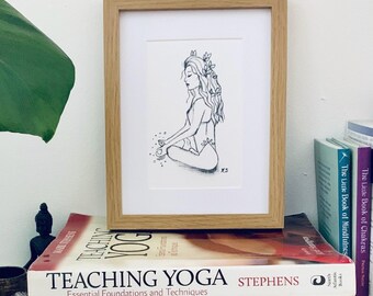 May include: A framed black and white line art drawing of a person sitting in a cross-legged meditation pose with leaves in their hair. The artwork is displayed in a light wood frame and rests on a stack of books titled Teaching Yoga and The Heart of Yoga.