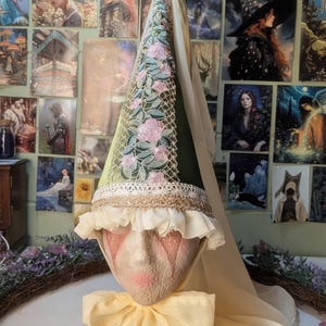May include: A whimsical, handcrafted doll head with a conical hat. The hat is green with pink floral embroidery and a white lace trim. A cream-colored ruffle and bow adorn the doll's face and neck, creating a fairytale aesthetic.